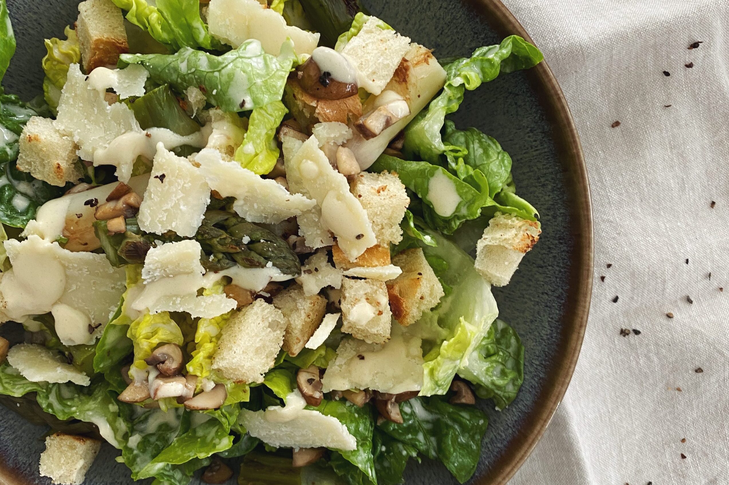 Caesars Salad mit Spargeln, gebratenen Pilzen, Brotwürfeln und hausgemachtem Dressing in einer blau-braunen Schüssel angerichtet.