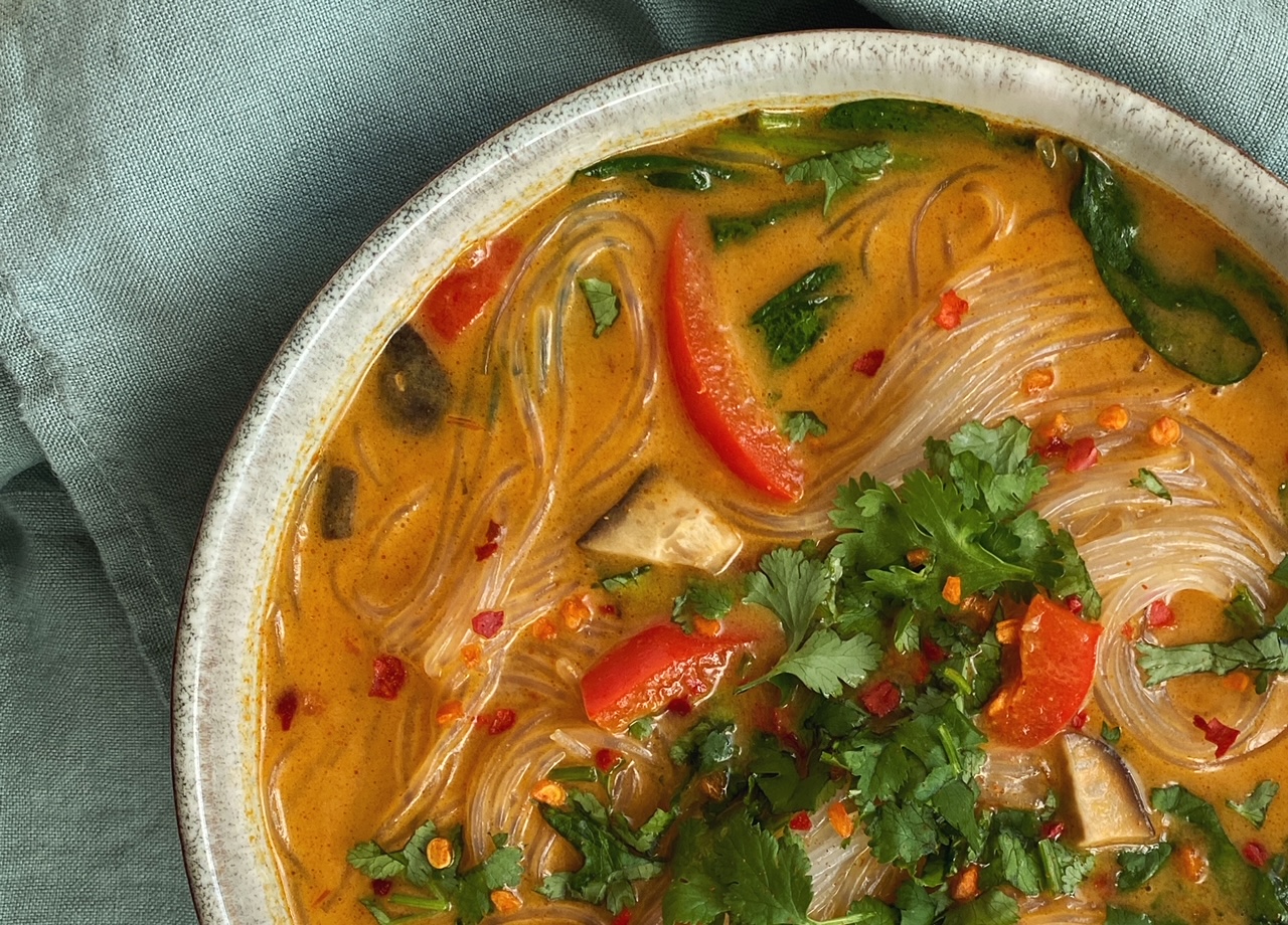 Rote Curry-Suppe mit Gemüse und Glasnudeln - von oben fotografiert auf einem türkisen Leinentuch. Mit Koriander und Chiliflocken garniert.
