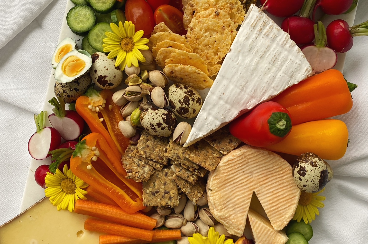 Frühlingshafte Käseplatte (Cheeseboard) von oben fotografiert auf einer wiessen Tischdecke. Der Käse wird mit buntem Gemüse, Eiern und Krackern ergänzt. Als Deko sind essbare gelbe Blüten auf dem Essen verteilt.