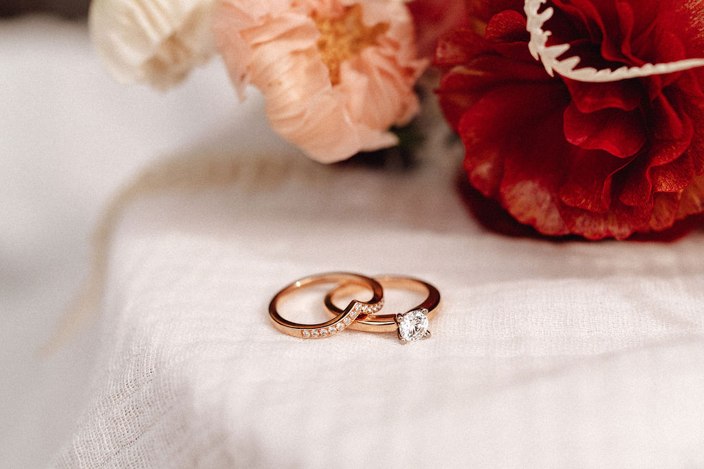 Zwei Ringe in geldgolb mit Diamanten von Sonja Denier liegen im Vordergrund des Bildes übereinander. Im Hintergrund sind Blumen aus dem Brautstrauss vom Blumenladen Florium zu sehen. Das Bild wurde von Cate Brodersen fotografiert