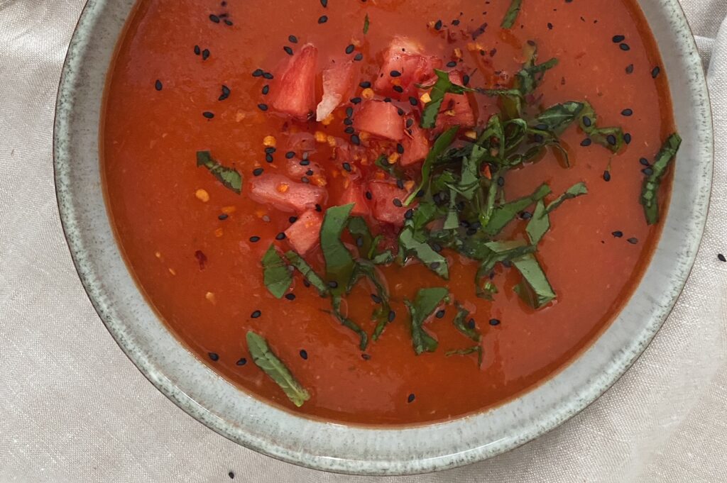 Erfirschende Gazpacho mit Wassermelone, von oben auf einem beigen Leinentuch fotografiert. Die Suppe wurde in einem graublauen Teller angerichtet und mit Wassermelonenstücken, Basilikum und schwarzem Sesam garniert